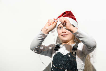Defocused foreground on child holding Christmas garland of snowflakes in his hands in front of his face on white background. Little girl in red hat of Santa Claus. Soft selective focus Merry Christmasの写真素材