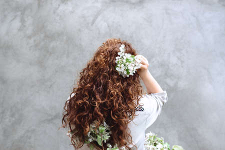 Back view of young curly brunette girl with long healthy hair spring flowers in hand background gray textured concrete wall. Inspiration of spring and summer. Concept of perfume, cosmetics. Copy Spaceの写真素材