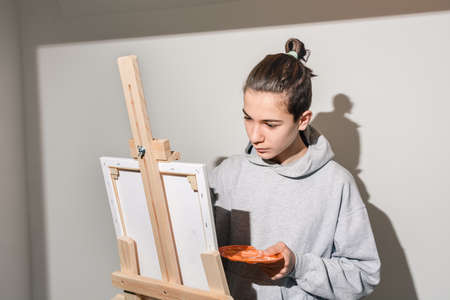 Teen artist intently paints picture on easel and holds brush. Child holds an orange palette in studio. Direct view, harsh light and uniform white wall. Guy in casual clothes. Copy Space, Soft focusの写真素材