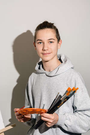 Portrait of a teenage artist holding an orange palette and many brushes in his hands on a background of white wall in the studio. Direct view, sharp light. The guy in casual clothes. Copy space.の写真素材