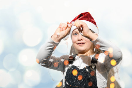 Defocused foreground on child holding a Christmas garland of snowflakes in front of his face on background of shimmering bluish-white wall. Little girl in red hat of Santa Claus. Soft selective focusの写真素材