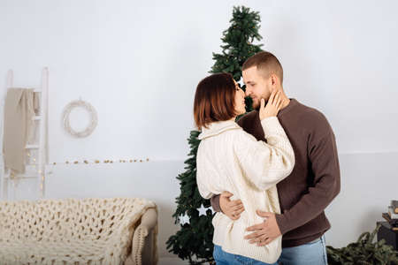 Beautiful young stylish couple, man and woman, in knitted sweaters white and brown in each others arms on the background of the Christmas tree celebrate Christmas at home. Scandinavian minimalism.の写真素材