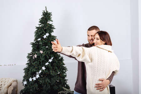 Cheerful young stylish couple, man and woman dancing, in knitted sweaters white and brown in each others arms on background of Christmas tree celebrating Christmas at home. Scandinavian minimalism.の写真素材