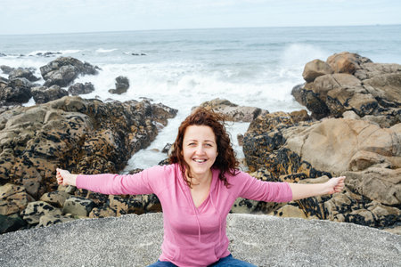 Young happy smiling girl sitting on the rocky shore of ocean and looking at camera on background of blue water hot sunny day. Woman in pink jacket. Tourist vacation. Porto beach, Portugal. Copy spaceの写真素材