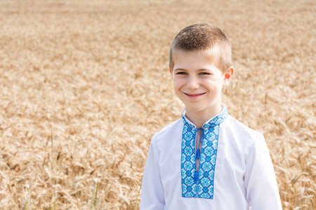 Soft selective focus of a schoolboy boy in an embroidered jacket on an agro-industrial field of a wheat field on a sunny day. The child smiles sincerely. Independence Day, Constitution of Ukraine.の写真素材