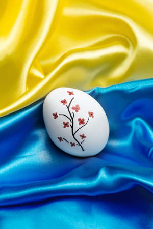 Vertical photo White Easter egg on the blue and yellow State Silk Flag of the Independence of Ukraine. A tree with red flowers. Happy Independence and Constitution Day. Happy Easter and holidays.の写真素材