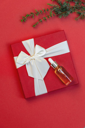 Mockup of glass dropper bottle with essential oil on gift box and Christmas tree branch on side on red background. Eco cream, cosmetics. Skin care product front view, copy space. soft selective focusの写真素材