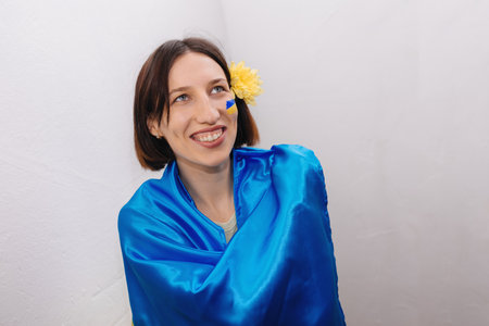 The flag of Ukraine is painted on girls cheek with blue and yellow paint. Flower in her hair. Woman smiles sincerely and looks away. Independence Day of Ukraine. Copy space.の写真素材