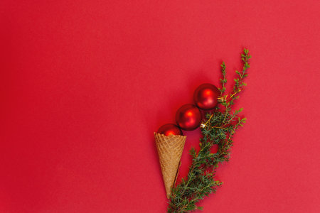 Three round toys in tasty waffle cone and next to it green branch of Christmas tree on red background. Concept of celebrating cold winter holidays in warmth. Top view, flat lay. copy spaceの写真素材