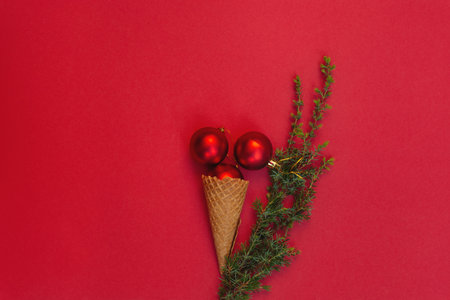Three round toys in waffle cone lie like delicious ice cream and next to it is green branch of Christmas tree on red background. Concept of celebrating cold winter holidays in warm. Top view, flat layの写真素材