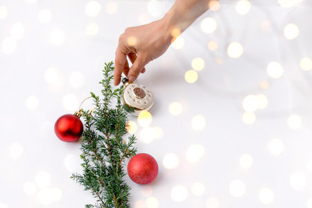 Female hand decorates Christmas tree branch with red round Christmas toys and clock on white background. Glowing yellow bokeh. Copy space for text. Selective focus on foreground.の写真素材
