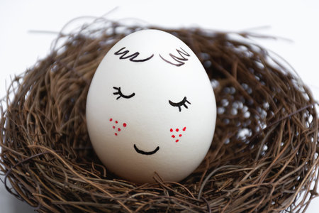 Close up of a beautiful white egg in a wicker nest on a white background. Cute face with red freckles. The view is straight. Spring postcard. Happy Easter.の写真素材