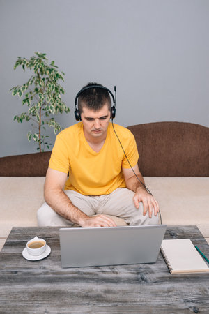 Young serious man in yellow t-shirt working on laptop from home and chatting online with client. Green plants in room. Concept of online learning and homework, freelancer. Work from home. Verticalの写真素材