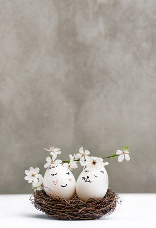 Happy Easter. White Easter eggs in wicker wooden nest on white table on gray background. view is straight. Blossoming branch of cherry with white flowers. Stylish spring template with copy spaceの写真素材