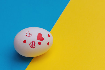 White Easter egg with various red hearts on yellow-blue background. Concept of Ukrainian refugees in search of safety and love for Ukraine. copy spaceの写真素材