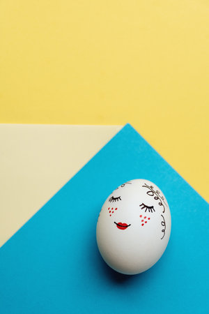 White Easter egg with cute drawn face lying on background of yellow and blue geometric paper. Concept of colors of Ukraine. copy spaceの写真素材