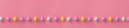Easter banner made of sweet colorful small eggs lying in one line on pale pink background. Festive sweets. Copy space for text. Lying flat. postcard.の写真素材
