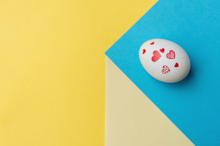 White Easter egg with various red hearts on yellow and blue geometric paper background. Concept of colors of Ukraine. copy spaceの写真素材