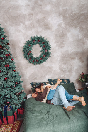 Happy couple hugging on a bed near beautifully decorated Christmas tree with gift boxes. Family in winter sweaters. copy spaceの写真素材