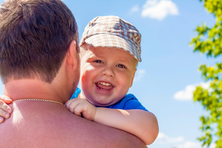 Portrait of a cute little boy. The boy sits in his father's arms and looks out from behind him. The...