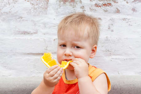 Portrait of child. Cute boy posing and eating a delicious orange. The emotions of a child. Portrait of child. Boy on the background of a brick wall.の写真素材