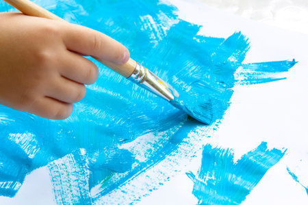 A child's hand holds a brush and paints paper with large strokes. Smears of blue paint on white...