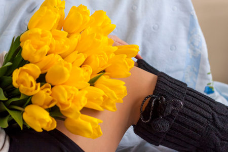 A bouquet of fresh yellow tulips lies on the lap of a young girl. The legs of a girl in knitted knee...