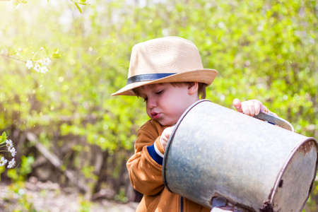 Cute little toddler boy in a hat and rubber boots is watering plants with a watering can in the garden. A charming little kid helping his parents grow vegetables. Active holidays with childrenの写真素材