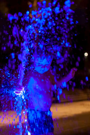 The child is frolicking in the fountain in the park, he is very happy. Have fun and enjoy the summer evening. Walking and recreation. Portrait of a happy child. Night shootingの写真素材