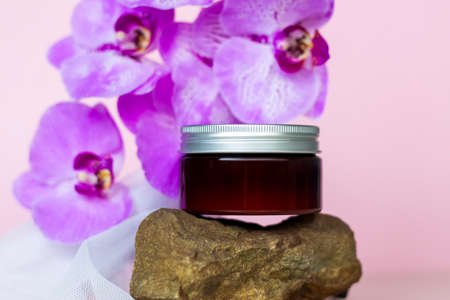 A brown jar of cream on a pink background stands on a large building stone with beautiful orchids next to it. Stylish appearance of the product, layout, personality. Banner, a place for the text.の写真素材