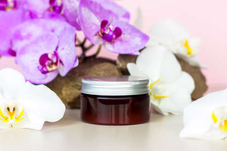 A brown jar of cream on a pink background stands on a large building stone with beautiful orchids next to it. Stylish appearance of the product, layout, personality. Banner, a place for the text.の写真素材