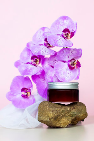 A brown jar of cream on a pink background stands on a large building stone with beautiful orchids next to it. Stylish appearance of the product, layout, personality. Banner, a place for the text.の写真素材