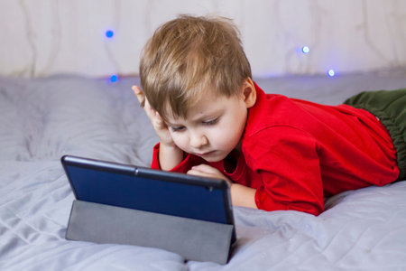 A cute boy is sitting on the bed and looking at a tablet. Funny smiling child. The concept of...