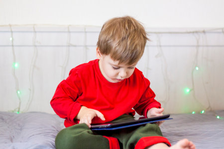 A cute boy is on the bed looking at a tablet. Online training, communication on the Internet. Funny smiling child. The concept of gadgets and children.の写真素材