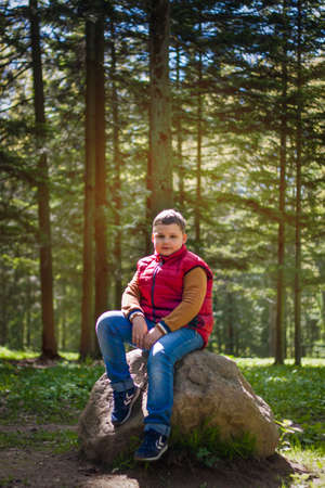 A young man in a sleeveless red jacket is sitting on a huge stone in a pine forest in spring. The magical light from the sun's rays falls behind the boy. Springの写真素材