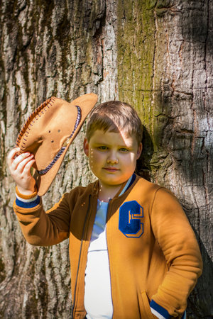 Cute boy posing in a cowboy hat in the woods by a tree. The sun's rays envelop the space. Interaction history for the book. Space for copying. selective focus.の写真素材