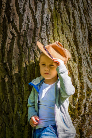 Cute boy posing in a cowboy hat in the woods by a tree. The sun's rays envelop the space. Interaction history for the book. Space for copying. selective focus.の写真素材