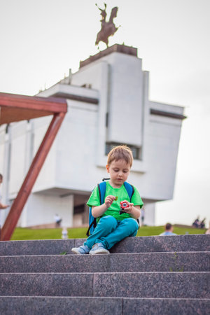 A boy is playing with a toy, sitting on the steps in the open air against the backdrop of skyscrapers and high-rise buildings. Journey. Lifestyle in the city. Center, streets.の写真素材