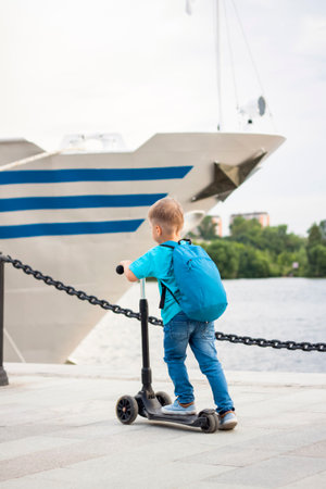 A boy on a scooter along the embankment of the city. Journey. Backpack on the back. The face expresses natural joyful emotions. Not staged photos from life.の写真素材