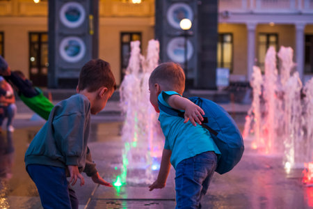 Children have fun frolicking in the fountains near the building in the park in the sunset light. Evening walk around the cityの写真素材