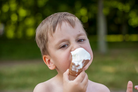 A cute blond boy appetizingly eats ice cream in the summer, sitting on the bank of the river. Cool off by the water. Funny expression. summer heatの写真素材