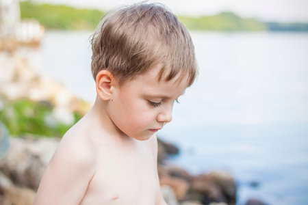 A cute blond boy experiences the joy of walking on a summer day by the river, sitting on the juicy grass. Funny expression. summer heat, a child without a T-shirt. Summerの写真素材