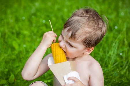 A cute blond boy appetizingly eats corn in the summer, sitting on the bank of the river on the juicy grass. Funny expression. summer heat, a child without a t-shirtの写真素材