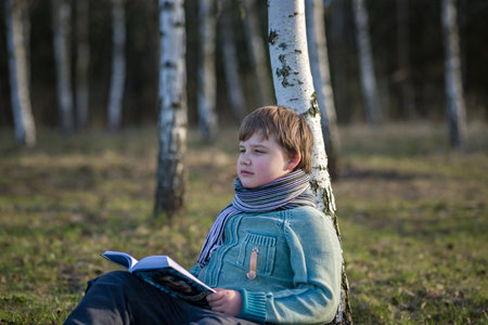 A handsome boy in a scarf is in a spring park, sitting under a birch tree, reading a book and enjoying his dreams. A happy child. Portraitの写真素材