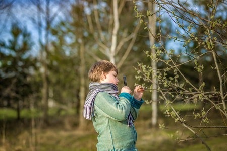 A funny boy walks through the spring park, examines the leaves with a large magnifying glass, looks...