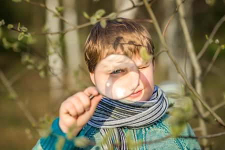 A funny boy walks through the spring park, examines the leaves with a large magnifying glass, looks for bugs and enjoys. A happy child. Portraitの写真素材