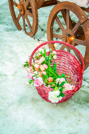 A wicker basket with beautiful flowers stands on the snow near the cart wheel. Flowers in a pink basketの写真素材
