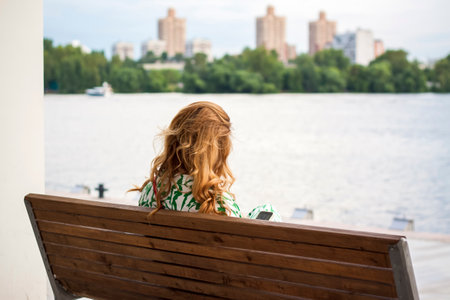 2022-07-17. Russia. Moscow. Northern River Station. A girl with long hair is sitting on a bench admiring the view of the river. view from the back. Other people are resting nearby. Moscow Riverのeditorial素材
