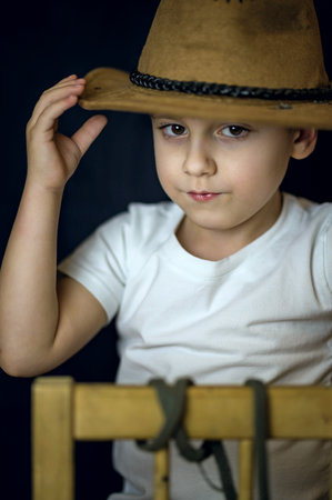 A concise portrait of a European boy. Portrait on a dark background of a boy in a cowboy hat and a white T-shirt. A strict frame.の写真素材