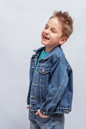 Cheerful young boy wearing denim jacket and jeans with tousled hair, standing with hands in pockets, smiling broadly, creating a playful and joyful expression against a plain backdropの写真素材
